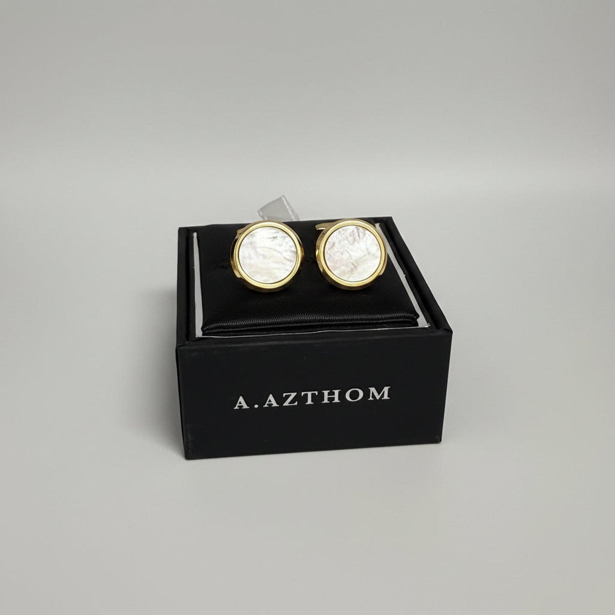A pair of round gold cufflinks with iridescent mother-of-pearl faces, presented in a black A. Azthom gift box.