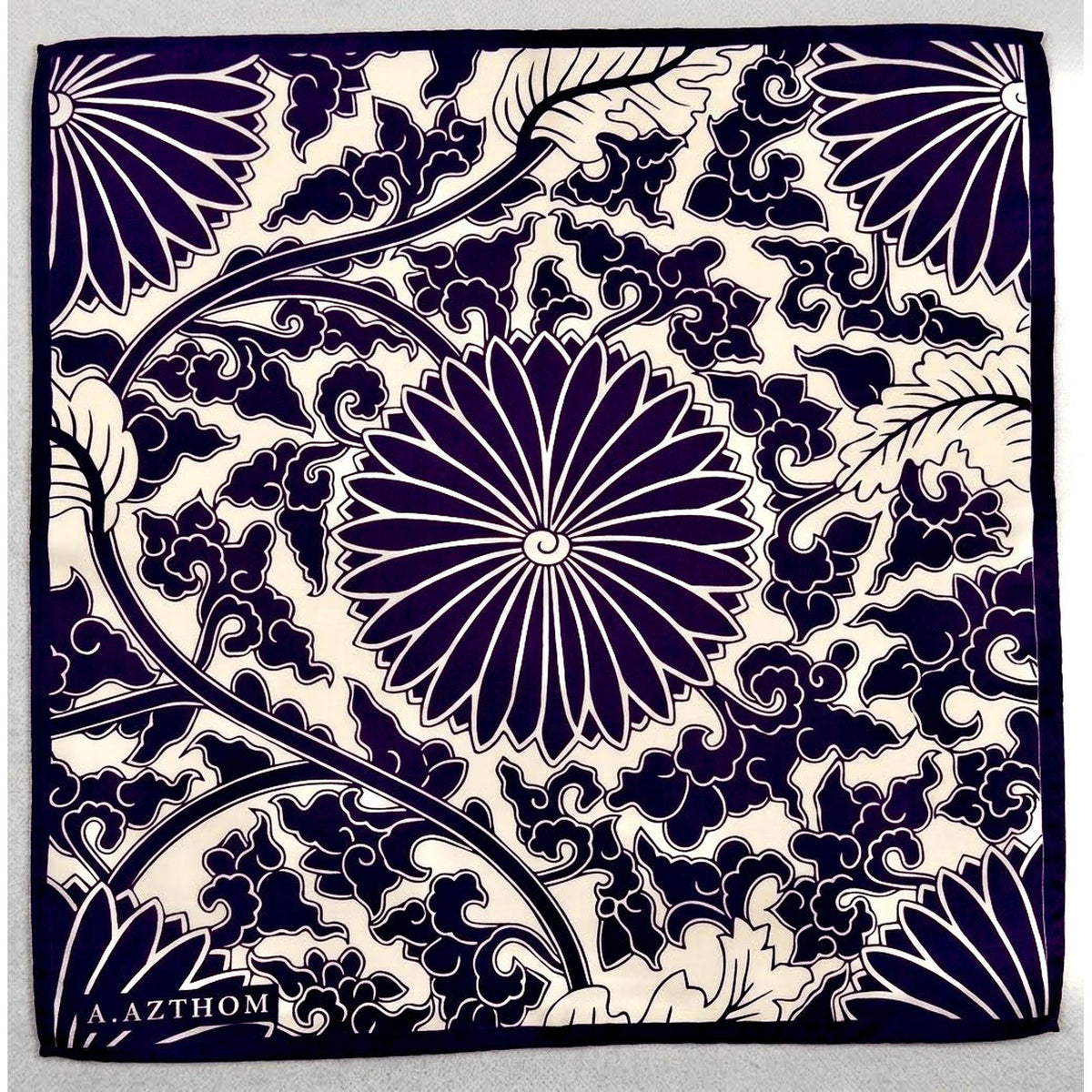 A square pocket squre  featuring a navy blue and cream floral pattern with a central chrysanthemum and swirling vine motifs. The brand name 'Azthom'