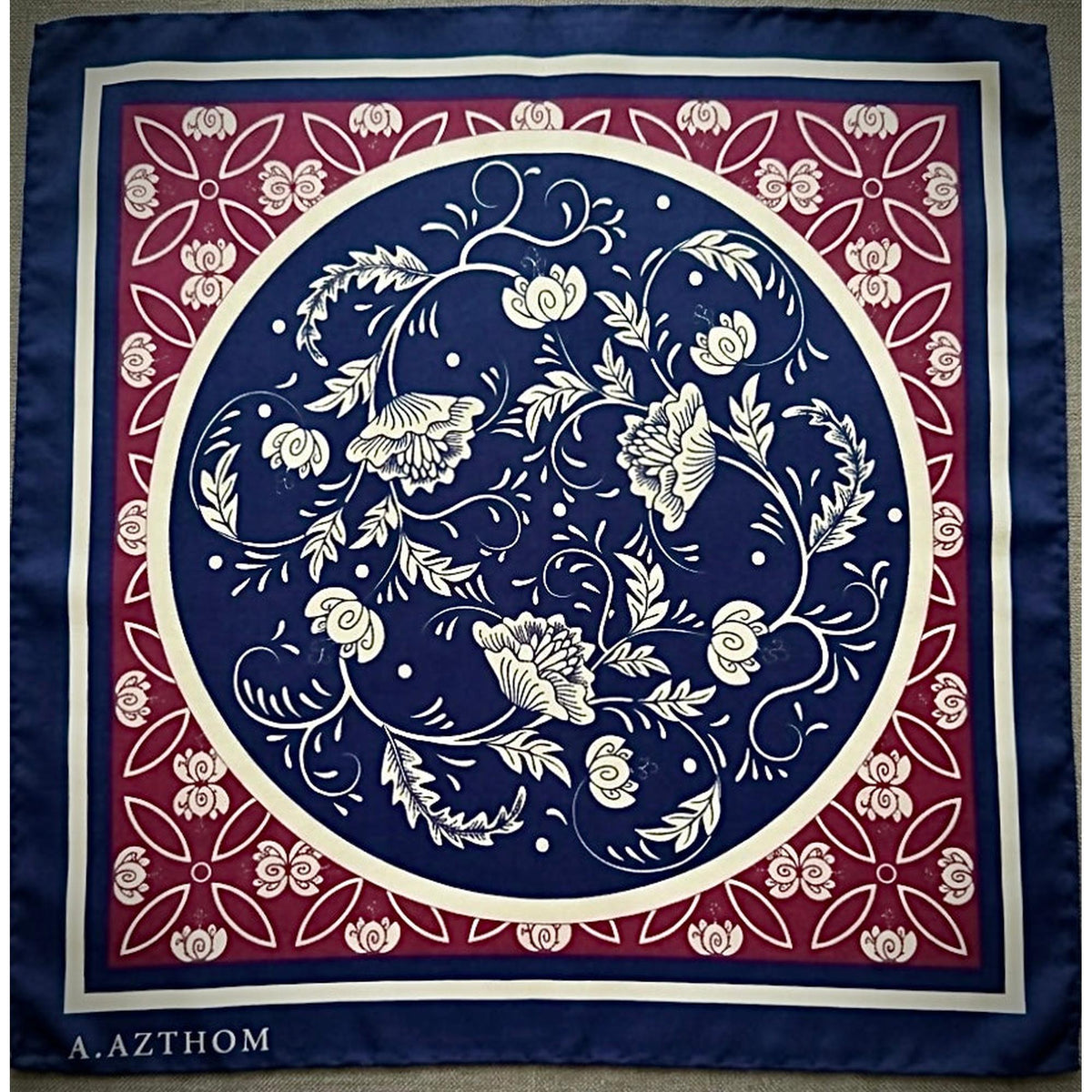A square silk scarf featuring a classic botanical pattern with a navy blue center, a red border, and cream accents. The brand name 'A.