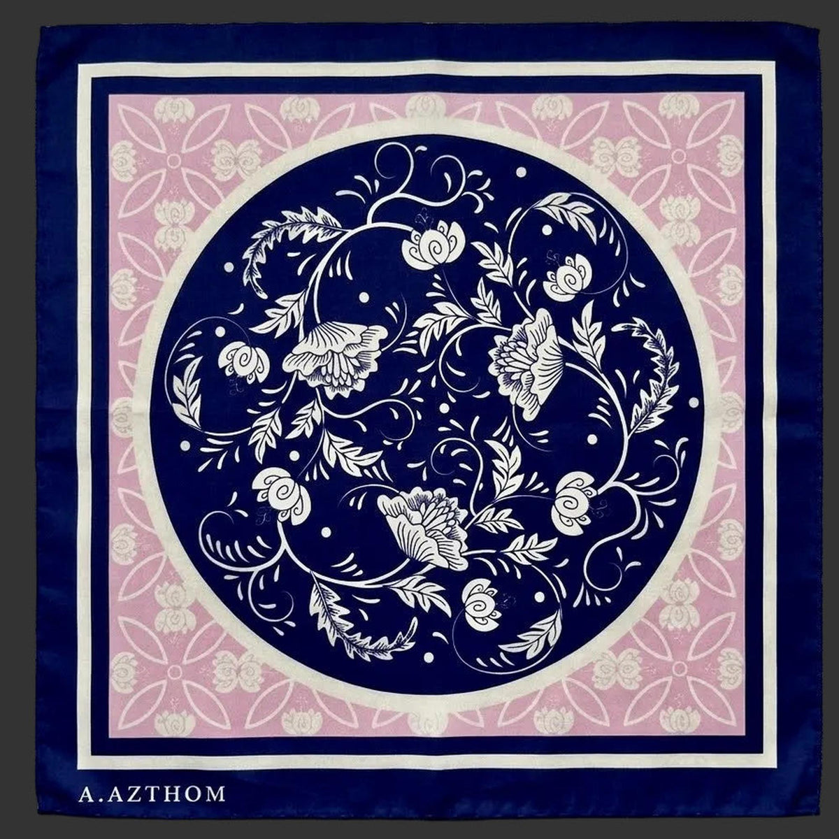 A square scarf laid flat on a gray textured fabric background. The scarf features a navy blue border, a central navy blue circle with white floral and vine motifs, a pink inner field with white floral patterning, and the brand name 'A.