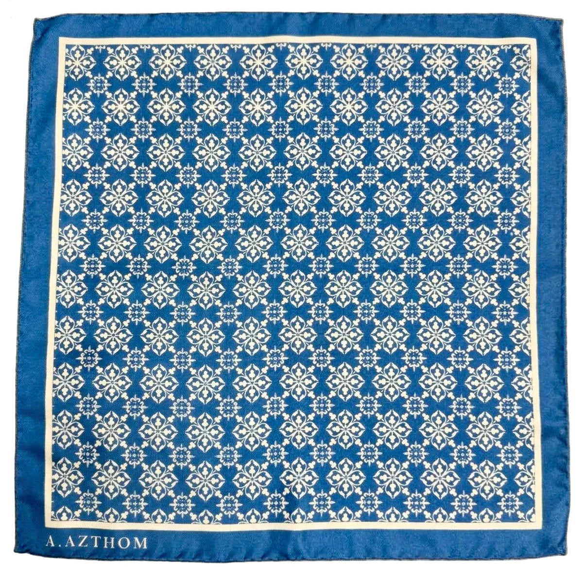 A square blue scarf with a white geometric damask pattern and a solid blue border, featuring the brand name 'A. AZTHOM' printed in white at the bottom left, laid flat on a gray carpet.